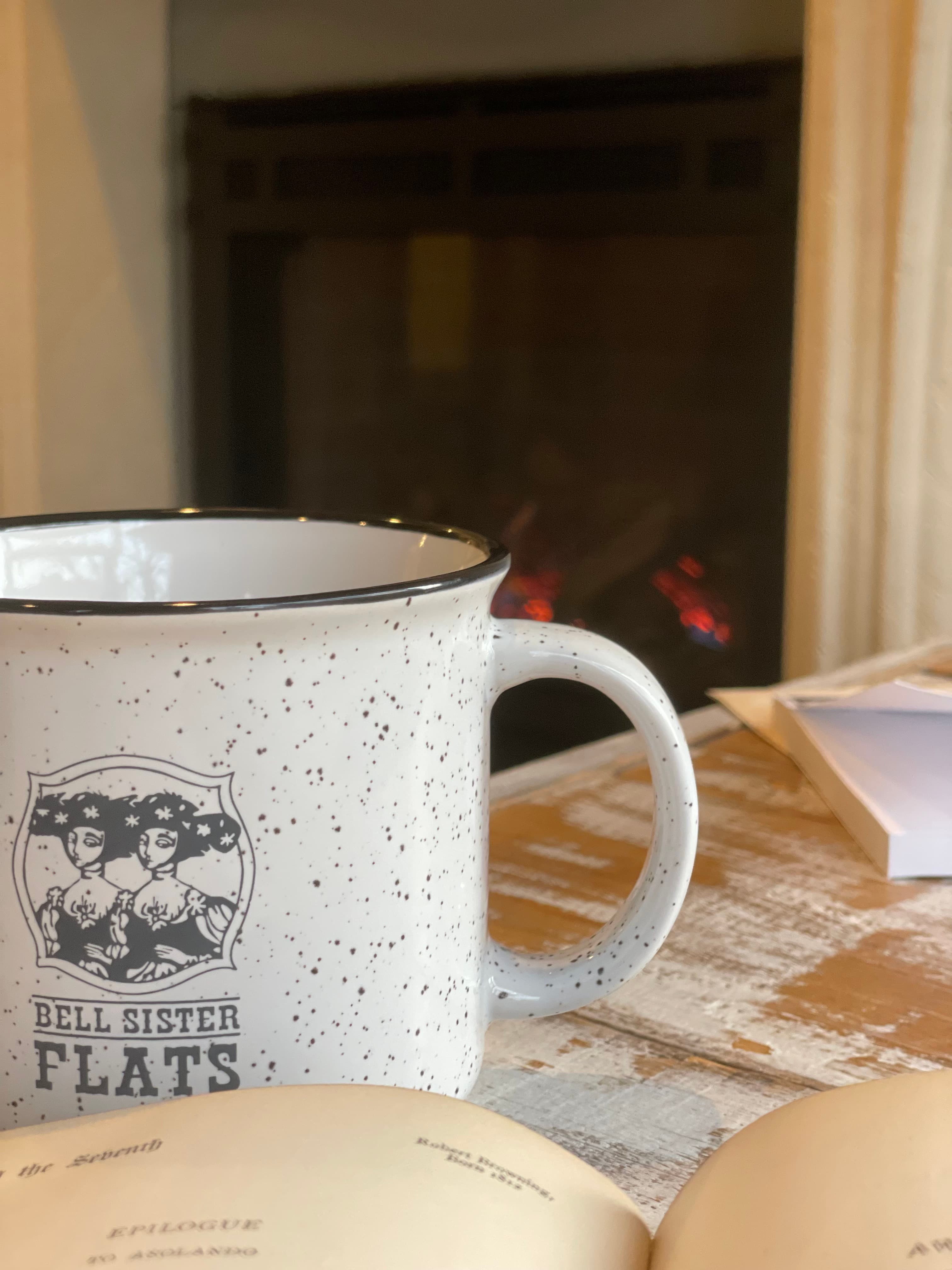 A speckled mug sits beside an open book on a wooden table, with a cozy fireplace in the background. A speckled mug sits beside an open book on a wooden table, with a cozy fireplace in the background.